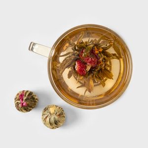 Glass teacup holds blooming tea, steeping in water; two unfurled tea balls rest nearby on a plain surface.