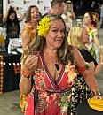 A woman in a floral dress holds a drink, standing among people in a festive indoor gathering with decorations and a bar area.