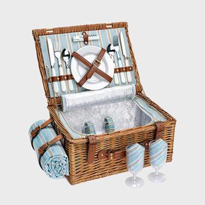 Wicker picnic basket holds utensils and plates, open in front of two glasses and a rolled blanket on a plain background.