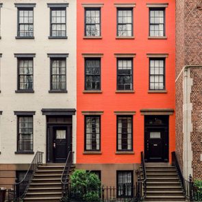 Three adjacent townhouses stand tall, displaying red, white, and brick facades. Each features multiple windows and stoops in a residential neighborhood.