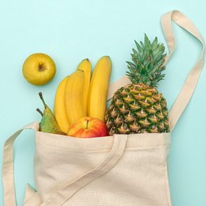 A canvas bag holds bananas, pineapple, pear, and apple, lying on a light blue surface with a yellow apple beside it.