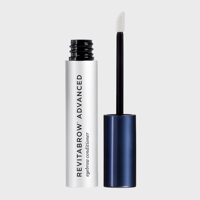 Tube of eyebrow conditioner stands open, applicator removed and upright, against a plain gray background. Text: 