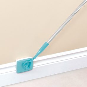 Mop scrubs white baseboard; turquoise cleaning head and handle, beige wall background.