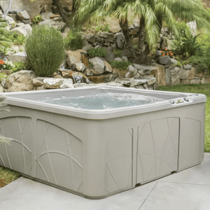 Hot tub bubbling, situated in a landscaped garden with rocks, plants, and palm trees surrounding it.