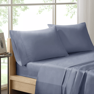Blue pillows rest on a neatly made bed by a large window, with a framed photo and books on a nearby table.