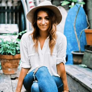 A woman sits relaxed, wearing a hat and white shirt, in a rustic outdoor setting with plants and wooden textures.