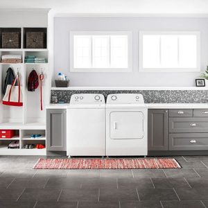 Washer and dryer sit idle in a well-organized laundry room with shelves, cabinets, and a red striped rug on a tiled floor.