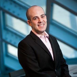 A person in a suit smiles while standing indoors against a backdrop of geometric window architecture.