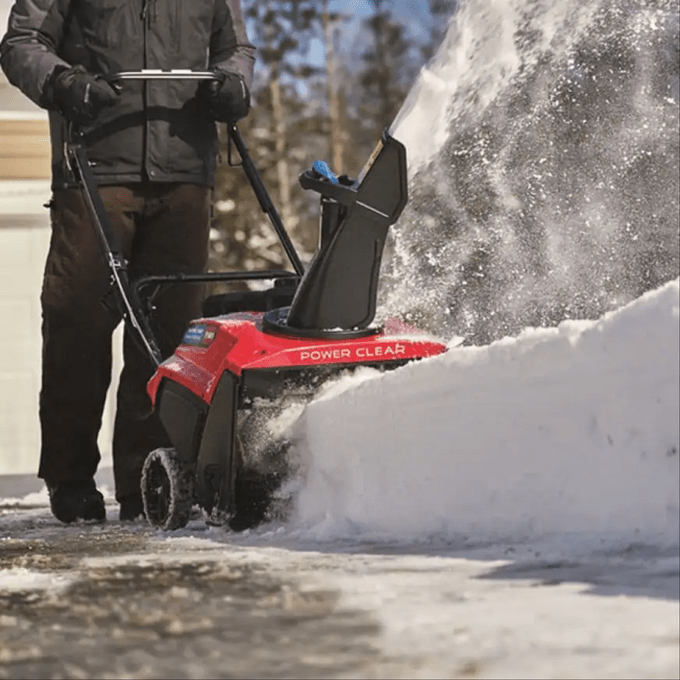 Snowblower clears snow on a driveway, operated by a person in winter clothing. Trees and a house are visible in the background.