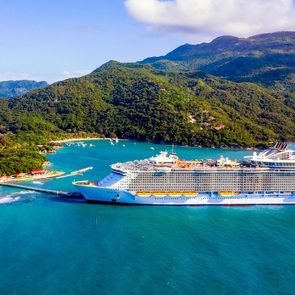 Cruise ship docked at tropical island port surrounded by lush green hills and turquoise water under a clear blue sky.