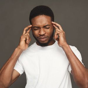 A person wearing a white shirt touches their temples, eyes closed, against a plain gray background.