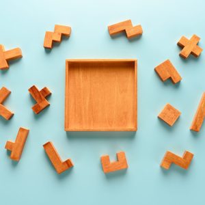 Wooden puzzle pieces surround an empty square wooden tray on a light blue background.