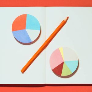 Colorful pie charts lie on an open notebook, with an orange pencil placed diagonally across, against a vibrant red background.