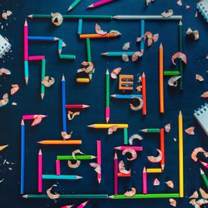 Colored pencils form a maze on a dark surface, surrounded by pencil shavings and notepads.