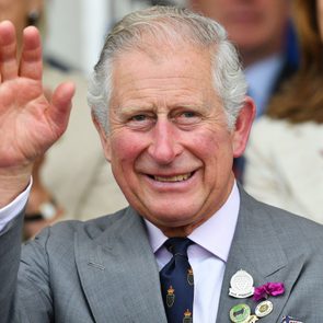 Man waves, smiling, in a gray suit with badges. Indoor setting, blurred people in background.