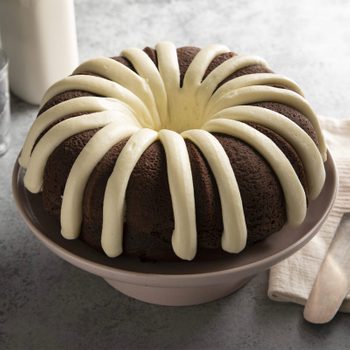 Bundt cake sits displaying white icing drizzled over chocolate surface, placed on a plate beside a folded napkin on a textured gray table.