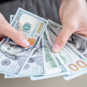 Hands hold and spread several 100-dollar bills, displaying currency prominently, with a neutral background.