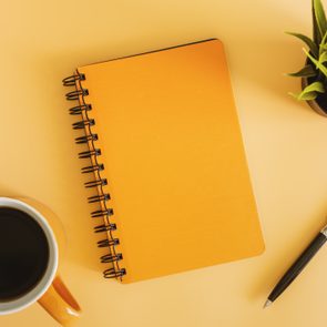 A bright orange notebook rests on a yellow surface beside a coffee mug, black pen, and green plant.