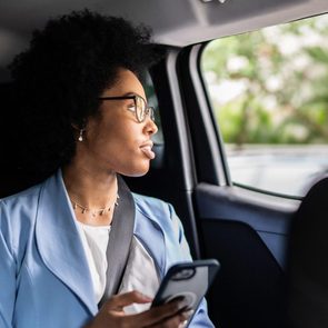 Person holds a smartphone, gazing thoughtfully out of a car window, surrounded by a blurred, green outdoor environment.