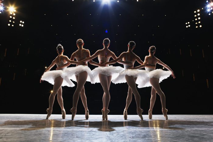 Five ballerinas en pointe on stage, arms around each other, rear view