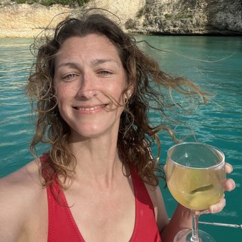 Person smiling, holding a wine glass; standing near turquoise water with rocky cliffs in the background.