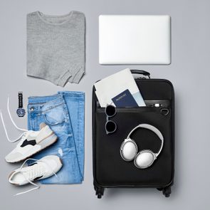 A gray sweater, jeans, watch, sneakers, laptop, and suitcase with accessories arranged neatly, suggesting preparation for travel.