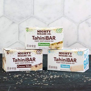 Three boxed TahiniBAR flavors displayed on a dark surface: Pistachio, Cocoa Nibs, Vanilla. White hexagonal tile background, scattered sesame seeds.