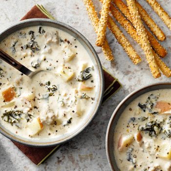 Bowls of creamy soup containing potatoes and greens, with a spoon in one bowl, surrounded by sesame breadsticks and brown napkins on a speckled surface.