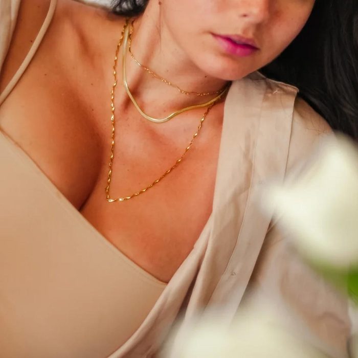 Necklaces layered on a woman's neck, resting, wearing a beige top, with blurred white flowers in the foreground.