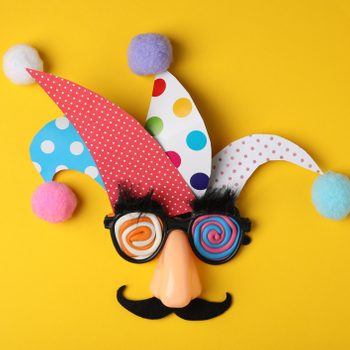 Clown accessories arranged creatively; jester hat, glasses with spiral eyes, mustache, and nose on yellow background.