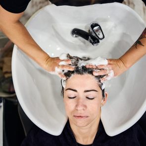 Person receives a scalp massage with shampoo in a salon, lying back into a white basin, surrounded by wooden floor and barber tools.