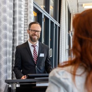 Receptionist smiling, engaging with a visitor; office lobby with patterned walls and glass doors in the background.