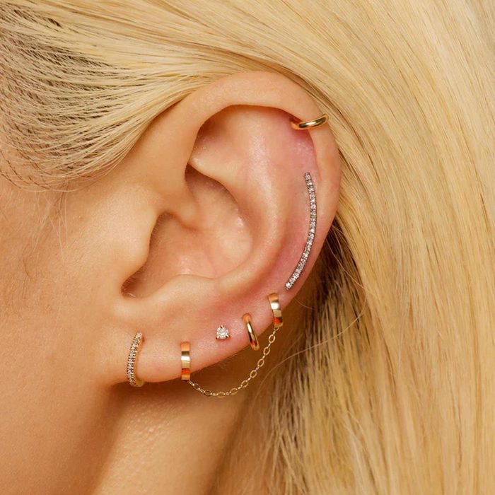 Earrings adorn an ear; multiple gold hoops and studs embellished with diamonds, some linked by a chain, amidst blonde hair.
