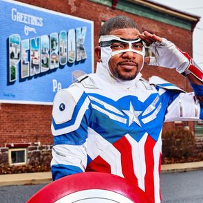 Person salutes wearing superhero costume with star emblem, standing in front of a brick wall with 