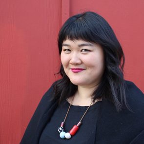 A woman smiles slightly, standing before a red background, wearing a black outfit and colorful bead necklace.