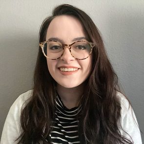 A person with glasses smiles against a plain, light-colored wall, wearing a black and white striped shirt and sweater.