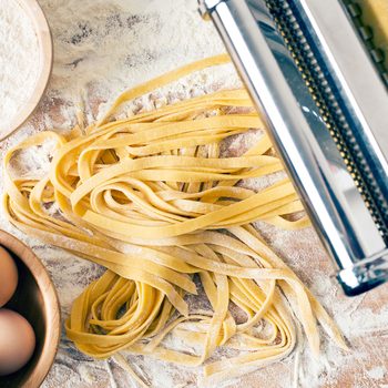 Fresh pasta lies on a floured wooden surface, emerging from a pasta machine, surrounded by bowls of flour and eggs.