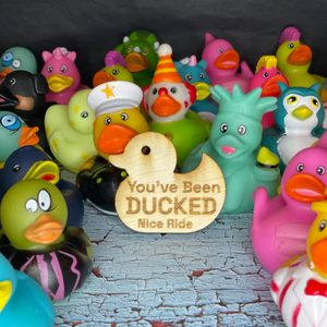 Rubber ducks are gathered, displaying colorful outfits, surrounded by a distressed, painted surface. A wooden tag reads, 
