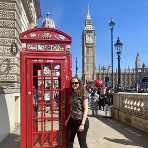 Red phone booth stands with a person nearby, Big Ben visible in the background amidst sunny, crowded London street.