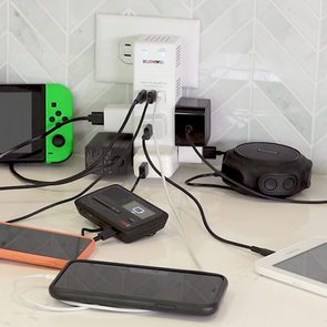 Power strip charging multiple devices on a countertop; includes phones, a game console, and a speaker. Walls have herringbone tile pattern.