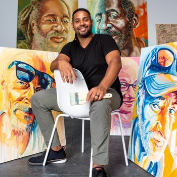 Artist holding a paintbrush sits on a chair, surrounded by vibrant, expressive portraits on canvases in a studio setting.