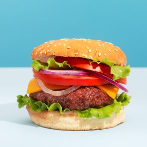Burger stacked with lettuce, cheese, tomato, and onions sits on a light surface, with a bright blue background.