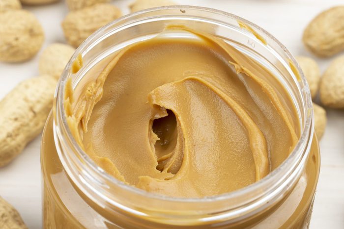 Jar of peanut butter on a white wooden table with peanuts scattered nearby. Space for text.