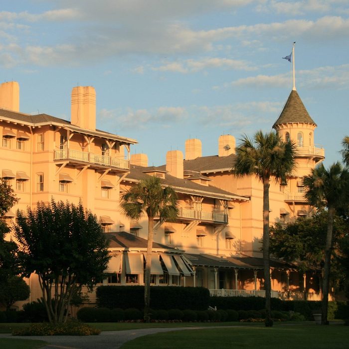 Large peach-colored building stands with a tower and balconies, surrounded by palm trees and greenery under a blue sky with scattered clouds.