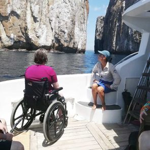 A person in a wheelchair faces majestic rock formations from a boat deck, accompanied by another person sitting on steps.