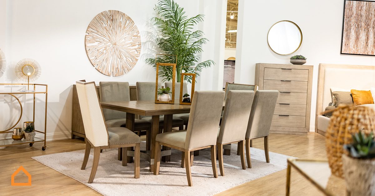 Dining table set with upholstered chairs arranged in a modern, stylish room; surrounded by decorative art pieces, plants, and wooden furniture, on hardwood flooring.