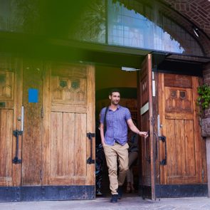 A person exits through wooden double doors of a brick building, with ivy on the wall, smiling and carrying a bag.