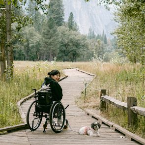 Person in wheelchair smiling on wooden path, next to small dog, surrounded by lush forest and grassy field.