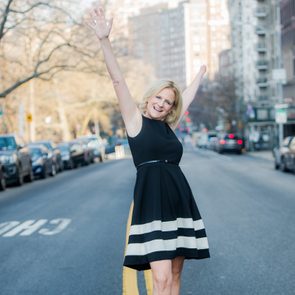 A woman in a black dress stands joyfully with raised arms on an urban street, surrounded by parked cars and buildings.