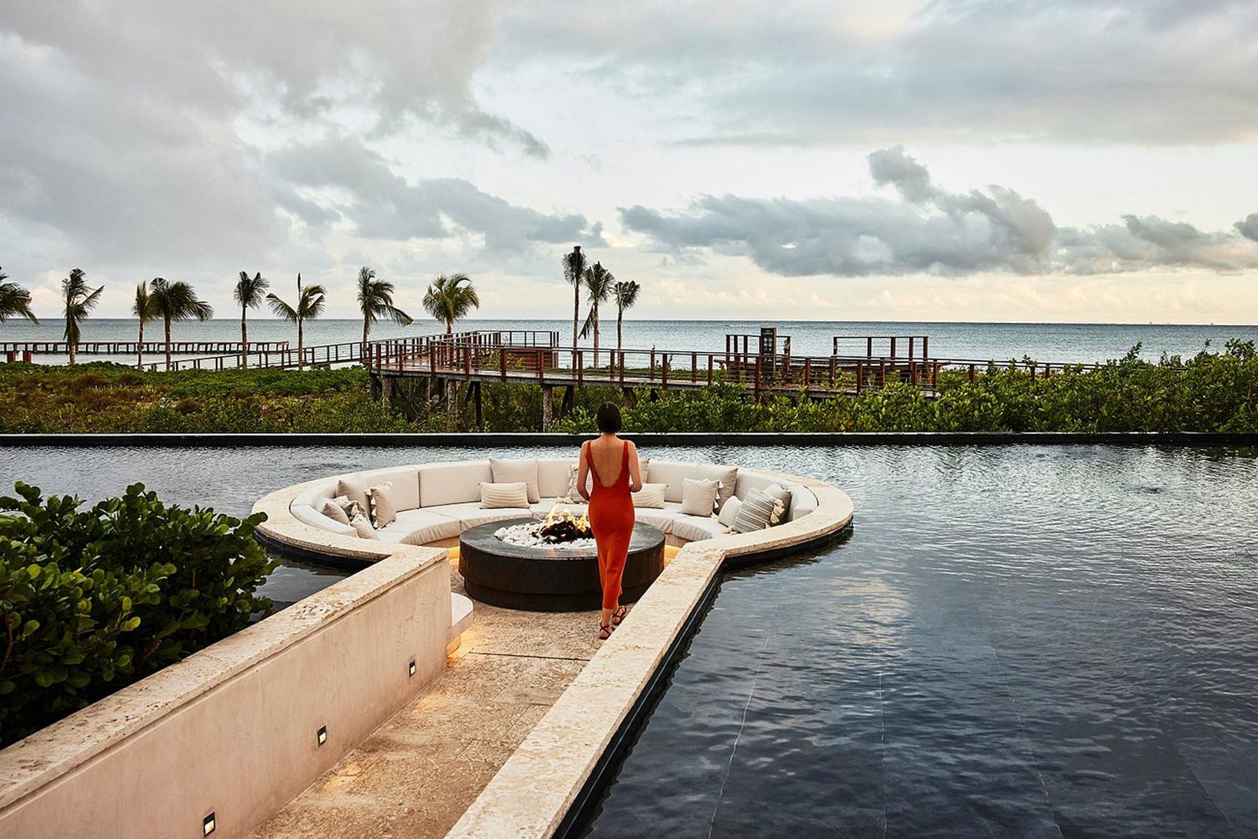 A Woman Standing by a Pool With a Fire Pit and a Body of Water